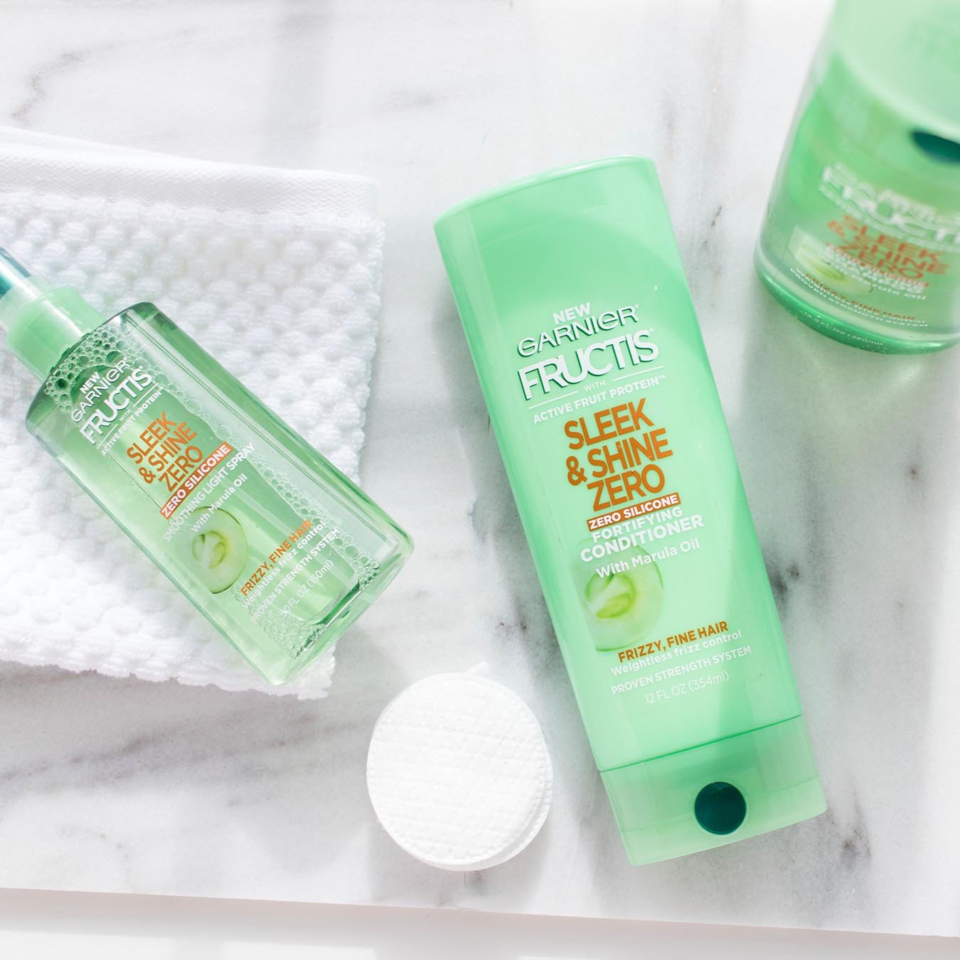 Garnier Fructis Sleek & Shine Zero Shampoo and Fructis Sleek & Shine Zero Conditioner on white marble and Fructis Sleek & Shine Smoothing Light Spray on a white wash cloth next to makeup removal pads.